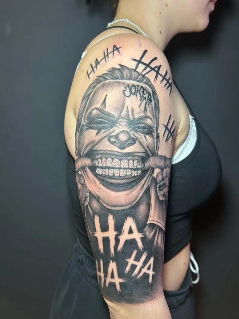 Black-&-Grey-Tattoo eines grinsenden Joker-Gesichts mit „HA HA“-Schriftzügen am Oberarm, gestochen von Cindy