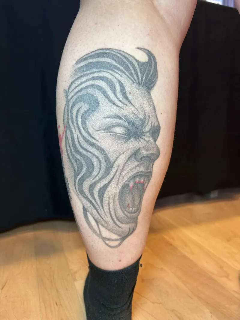 Black-&-Grey-Tattoo eines schreienden Gesichts mit wellenförmig verzerrter Haartolle an der Wade, gestochen von Cindy