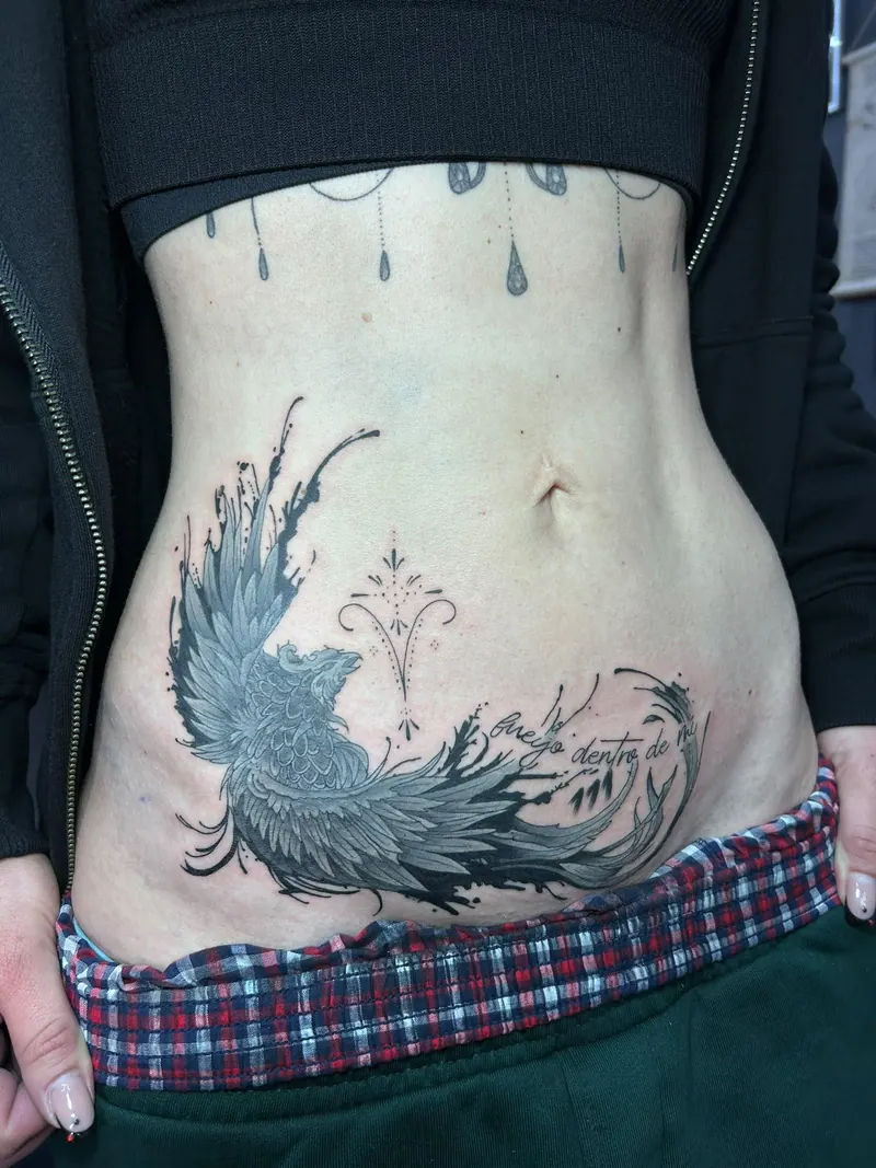 Black-&-Grey-Tattoo eines Phönix mit feinem Ornament und Schriftzug am Unterbauch, gestochen von Cindy