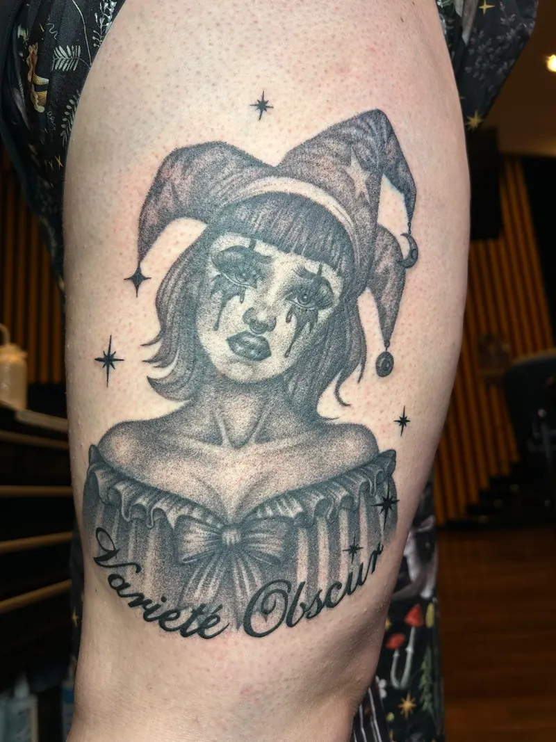 Black-&-Grey-Tattoo eines Pin-up-Portraits im Harlekin-Kostüm mit Schriftzug „Varieté Obscur“ am Oberarm, gestochen von Cindy