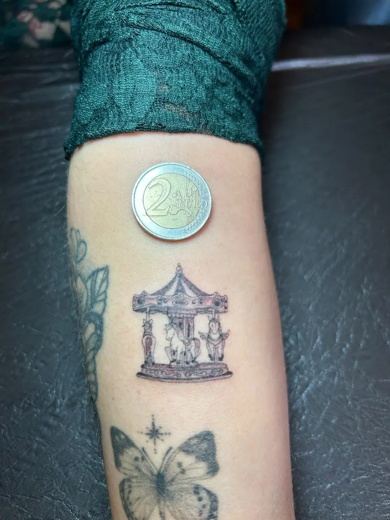 Kleines Black-&-Grey-Tattoo eines Karussells, neben einer 2-Euro-Münze als Größenvergleich, am Unterarm, gestochen von Cindy