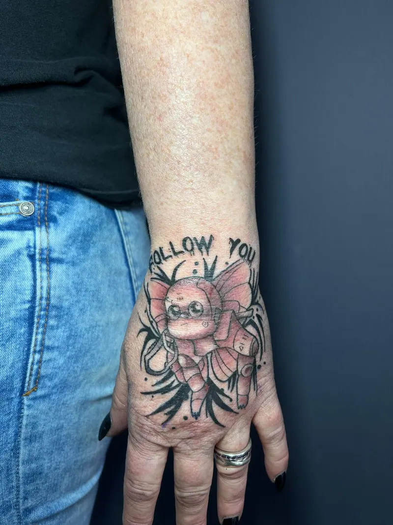 Farbiges illustratives Tattoo einer geflügelten Figur mit Schriftzug „Follow you“ auf dem Handrücken, gestochen von Cindy