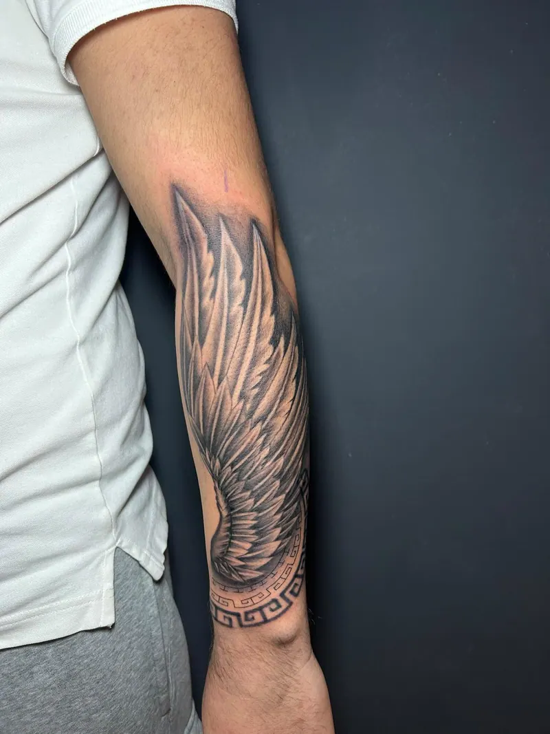 Black-&-Grey-Tattoo eines Flügels mit griechischem Mäanderband am Unterarm, gestochen von Cindy