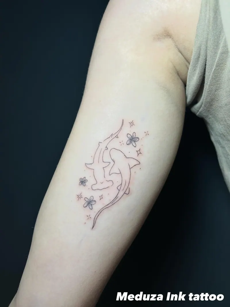 Fineline-Tattoo eines Hammerhais mit kleinen Blüten und Sternen am Oberarm, gestochen von Cindy