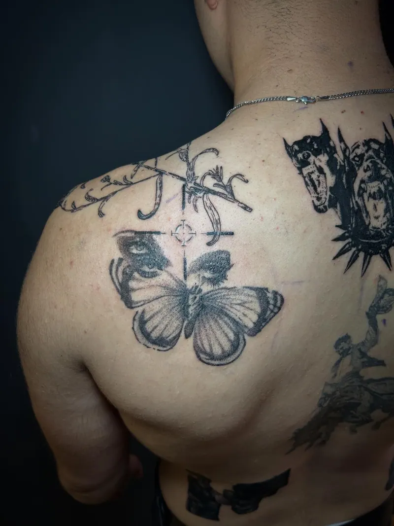 Black-&-Grey-Tattoo eines Schmetterlings mit Augen auf den Flügeln und Fadenkreuz am Schulterblatt, gestochen von Cindy