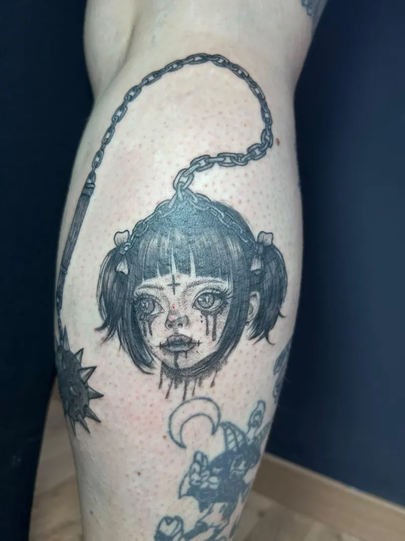Black-&-Grey-Tattoo eines weinenden Mädchenkopfs mit Zöpfen an einer Kette mit Morgenstern an der Wade, gestochen von Cindy