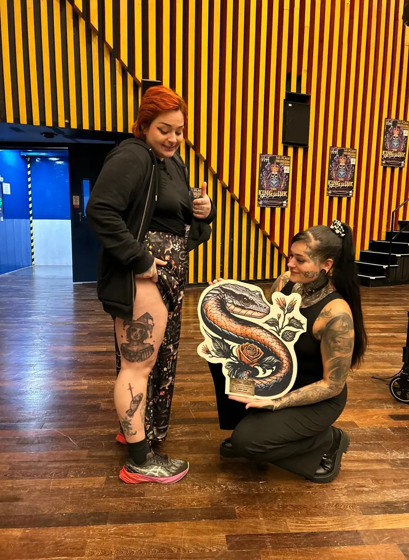 Cindy kniet mit Siegerschild neben ihrer Kundin, deren Bein-Tattoo prämiert wurde