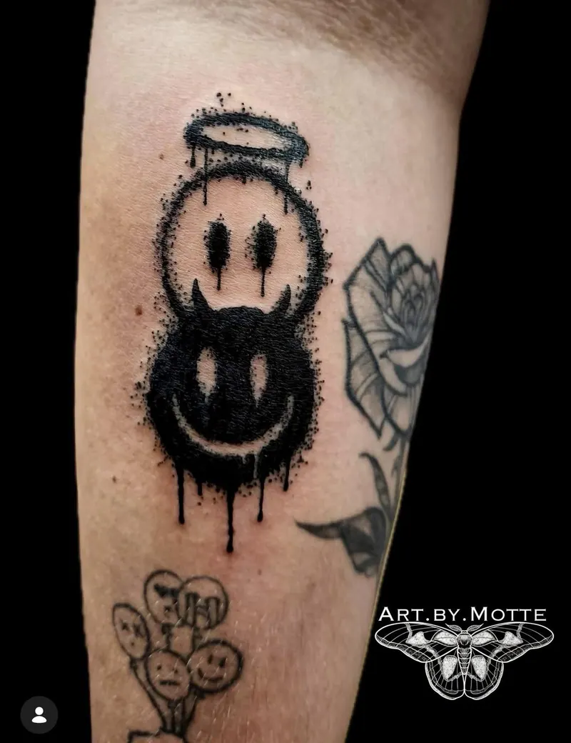 Blackwork-Tattoo eines Engel- und eines Teufel-Smileys mit Farbtropfen am Unterarm, gestochen von Motte