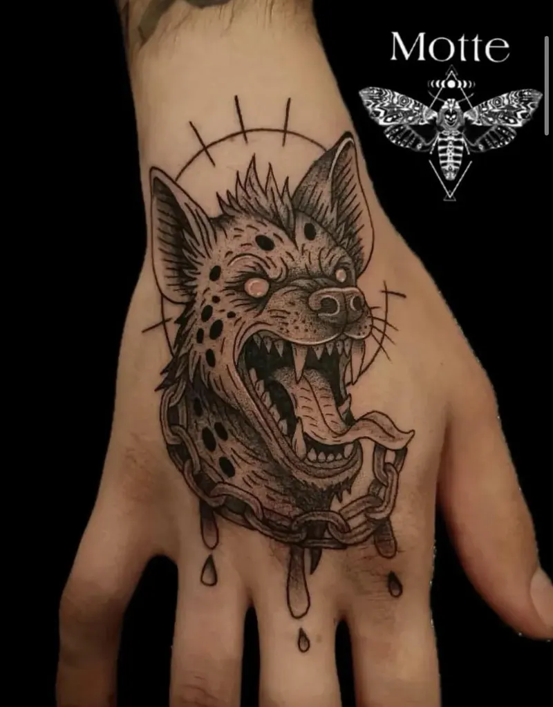 Blackwork-Tattoo eines Hyänenkopfs mit Kette und Halbkreis auf dem Handrücken, gestochen von Motte