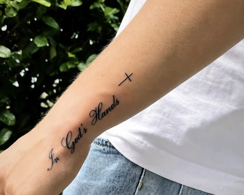 Lettering-Tattoo „In God’s Hands“ mit kleinem Kreuz am Unterarm, gestochen von Paul